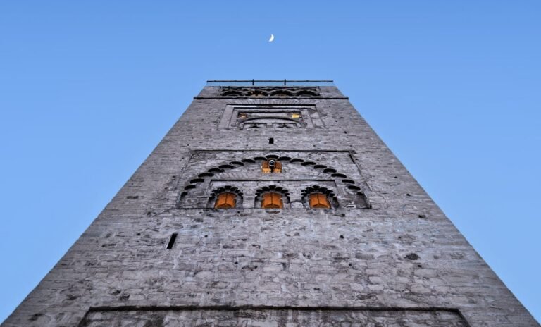 Low angle view of Koutoubia Mosque minaret under a crescent moon and clear sky in Marrakech.