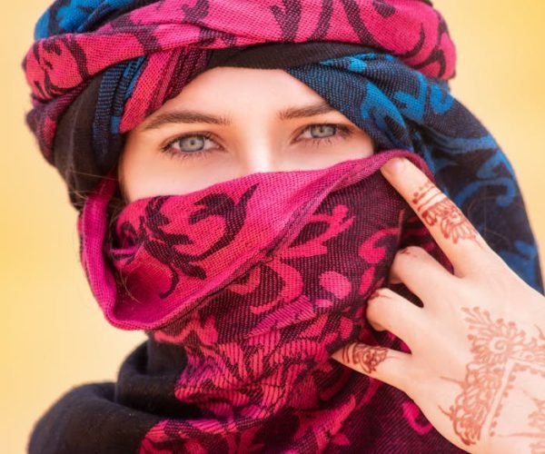 A woman in Merzouga, Morocco, wearing a colorful hijab and henna tattoo, standing in the desert.