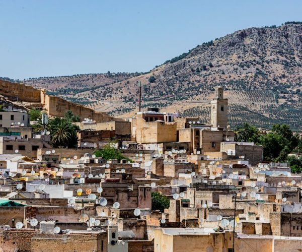 Explore the vibrant rooftops of Fes with satellite dishes, against a backdrop of Moroccan mountains.