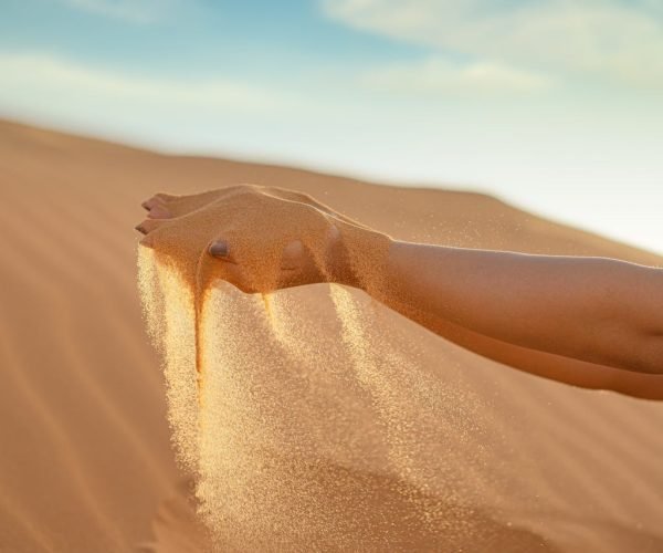 A hand releasing fine sand against the vast landscape of Merzouga Sand Dunes, Morocco.