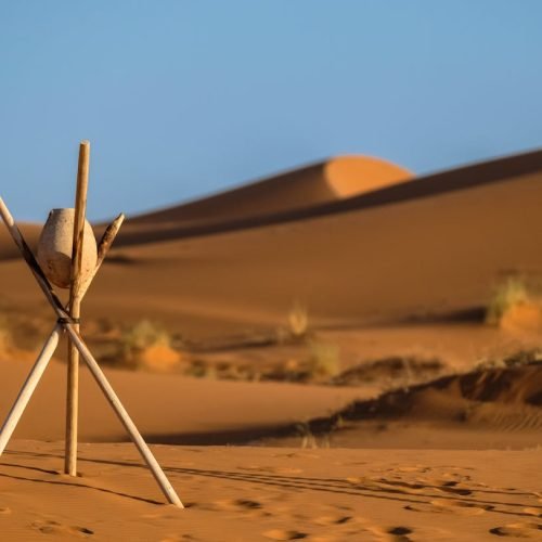 Peaceful desert landscape at Merzouga with sand dunes and wooden tripod under clear blue sky.